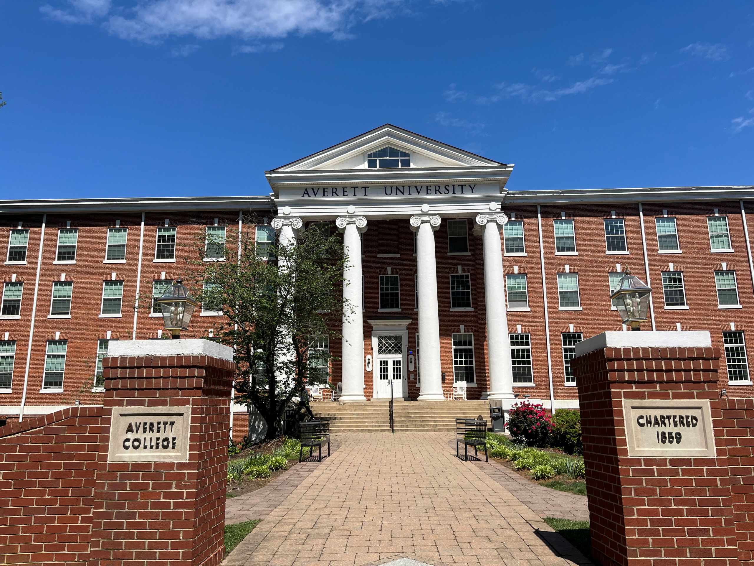 A brick gate stands outside a walkway to a large brick building with many windows and white columns, the main hall at Averett University in Danville.