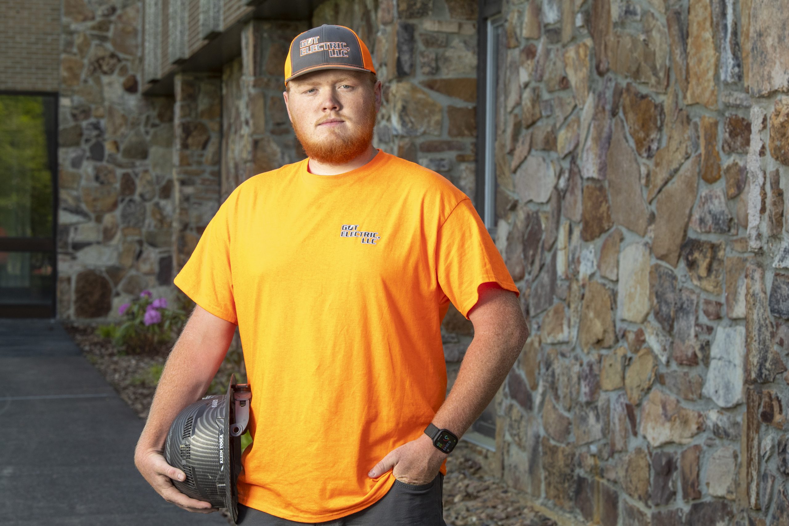 A young electrician in an orange t-shirt holds a hard hat at his hip while staring at the camera.