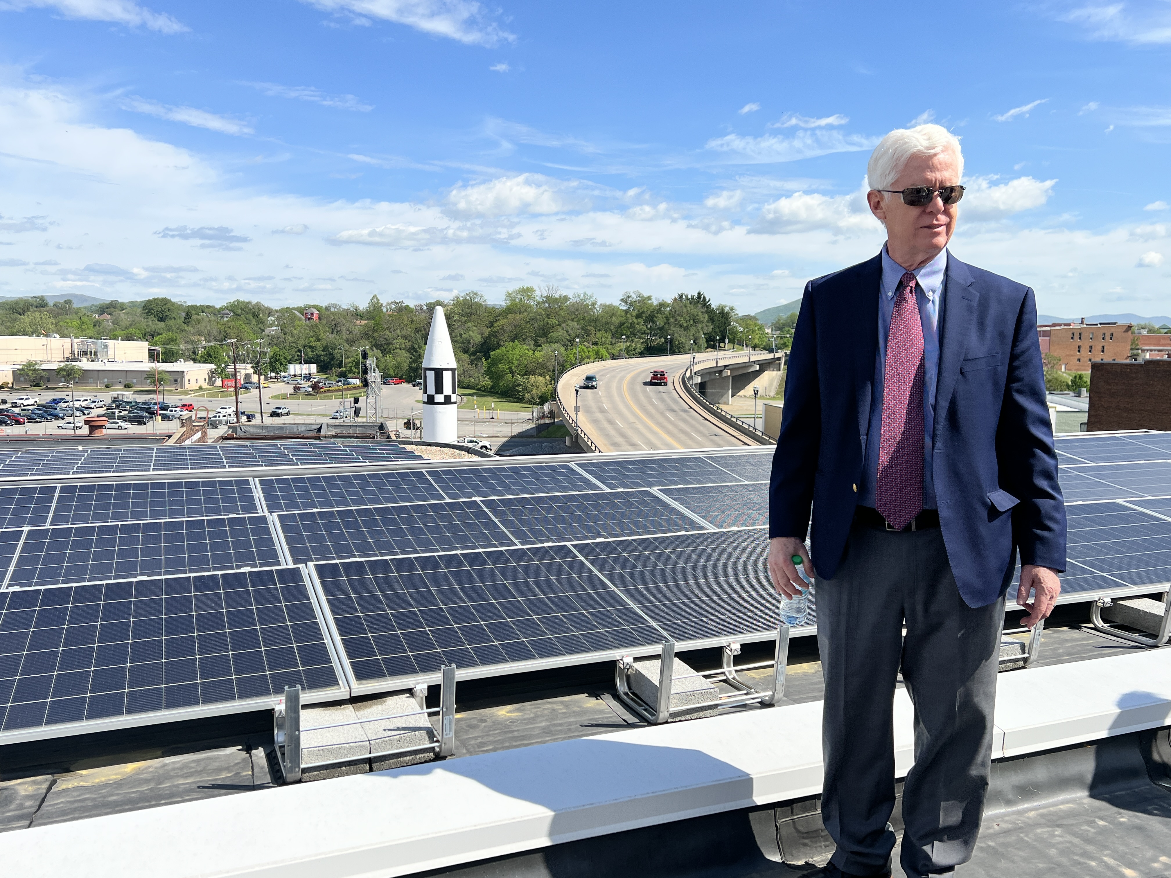 A man stands in front of a rooftop array of solar panels that overlooks a bridge and museum.