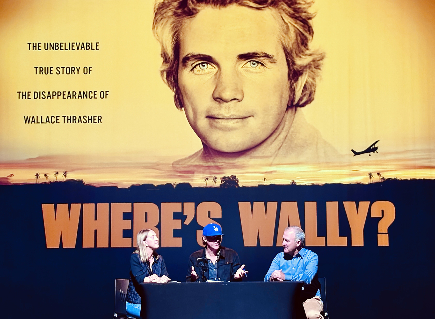 A woman and two men sit at a table on a stage under a giant movie poster that reads "Where's Wally?"