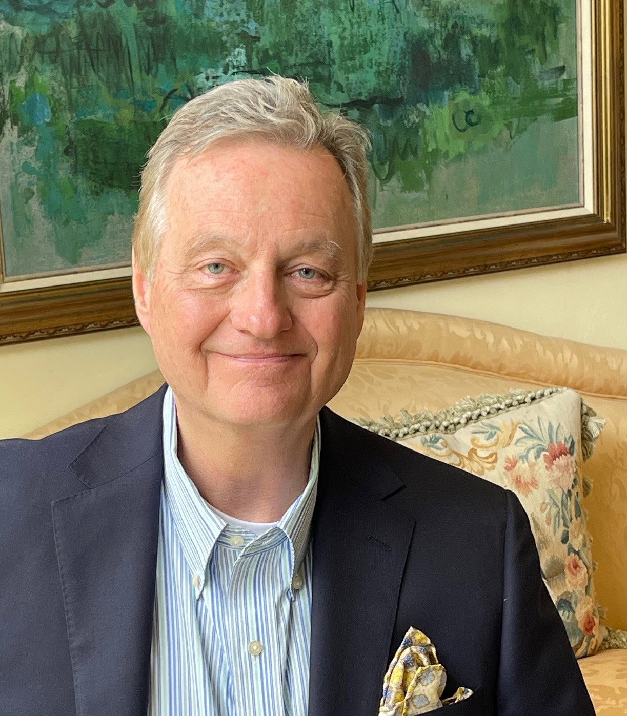 Portrait of Roanoke Blacksburg Technology Council member Russ Ellis, wearing a blue blazer with a yellow and blue-patterned handkerchief in the pocket and a thin-striped, button-down dress shirt. He is sitting on a couch, with a flower-patterned pillow and a painting behind him.