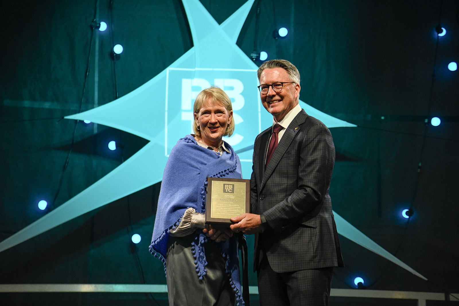 Carilion Clinic CEO Emeritus Nancy Howell Agee, wearing a blue shawl over a collared shirt and black pants, and Virginia Tech President Tim Sands, in a drak suit and burgundy tie, stand at the photo's center, holding Agee's Hall of Fame award from the Roanoke Blacksburg Technology Council. Behind them is a dark background, with light blue planet and moon representations, and a larger, star-shaped RBTC logo.
