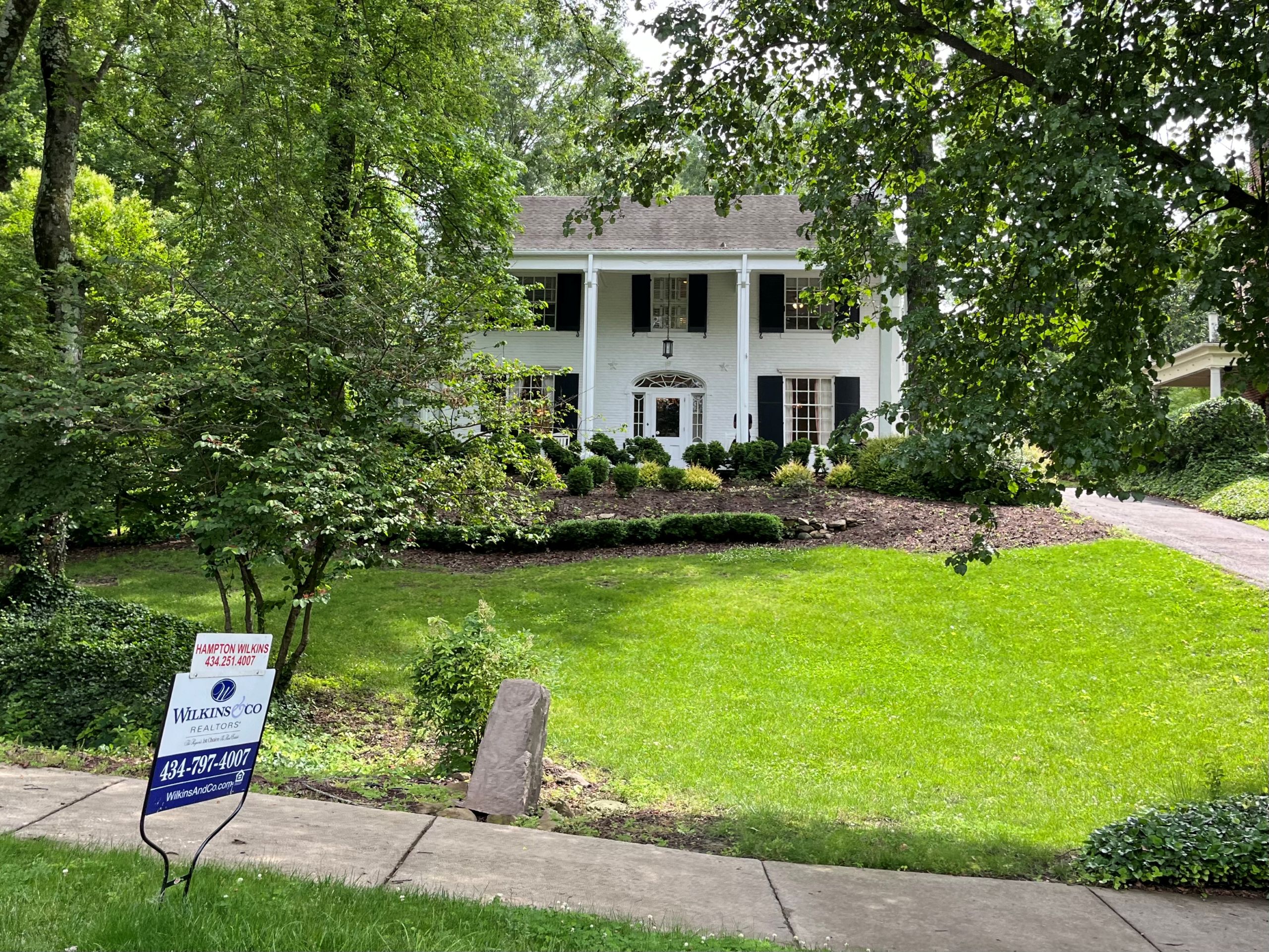 a large white house, the president's home for Averett University, stands behind a sidewalk where a for sale sign is posted