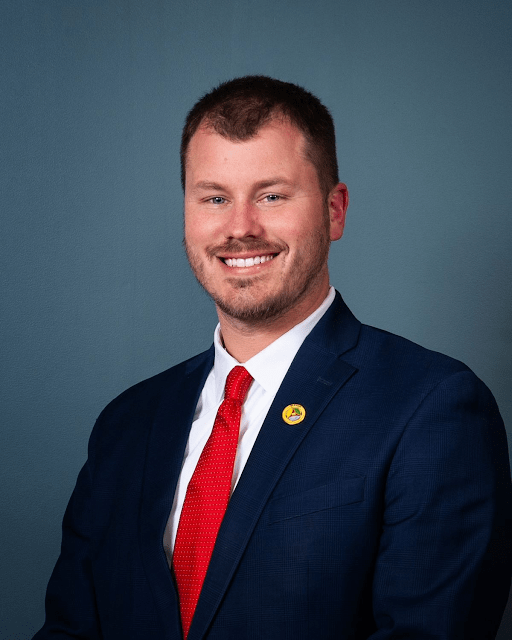 Rocky Mount Mayor Holland Perdue in a posed head shot for the town wears a dark suit and red tie, in front of a blue background.