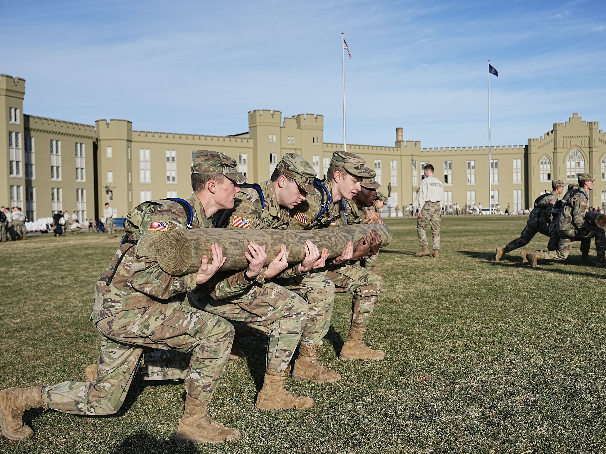 Four students wearing fatigues work together to carry a log across a grassy field in front of the VMI barracks.