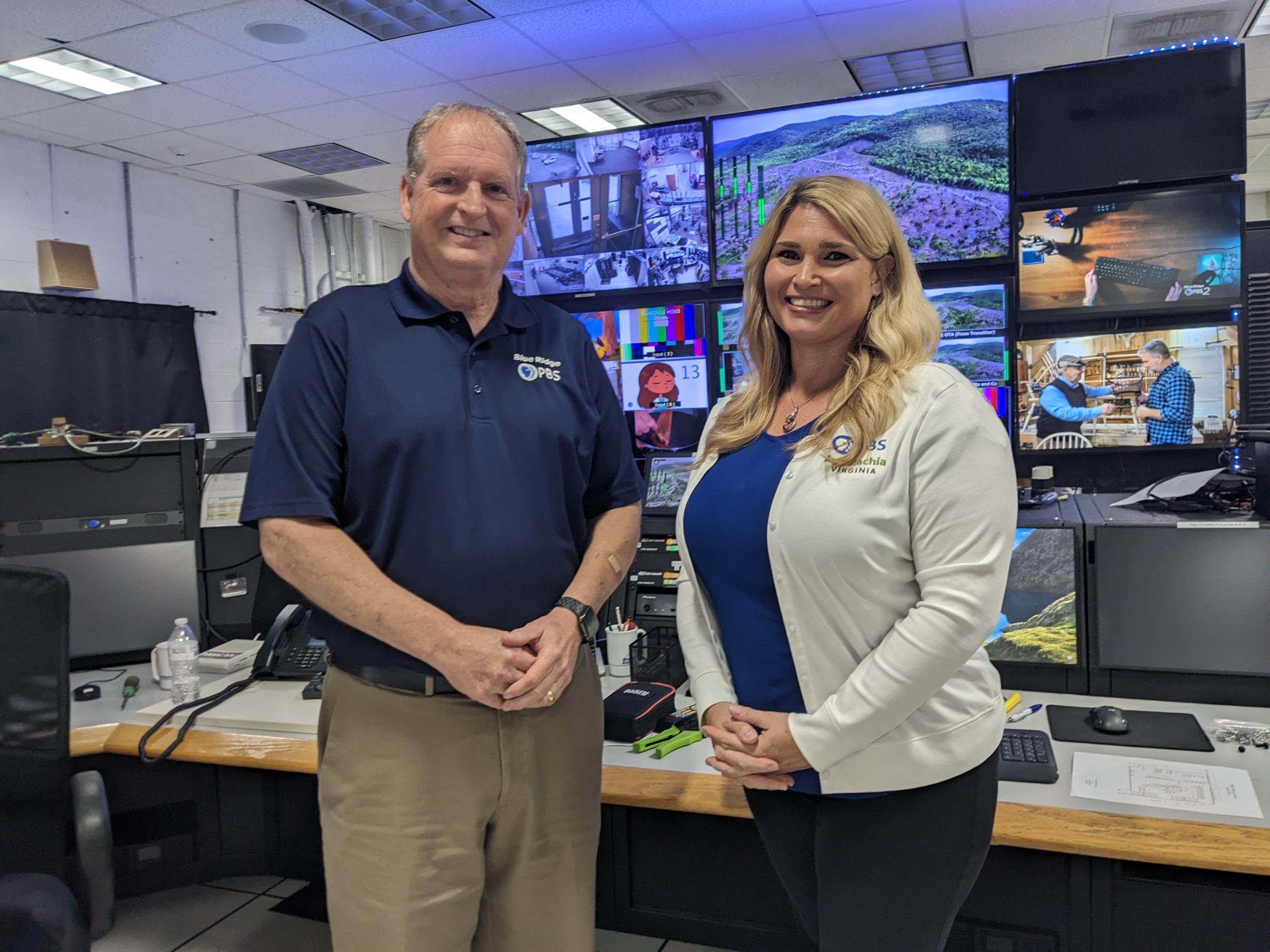 A man and a woman stand in front of a wall of monitors in a TV control room