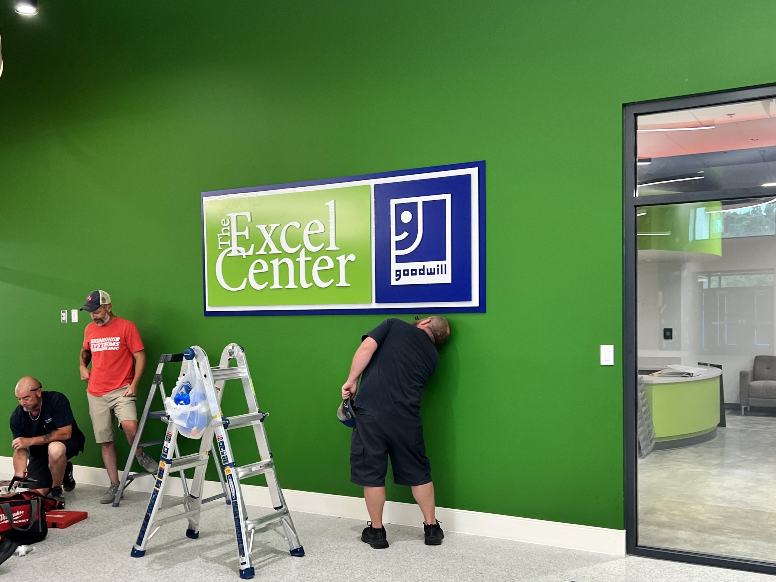 A man peers underneath a large sign hung on a green wall. It says "The Excel Center" and features a Goodwill logo. Two ladders can be seen, as well as a few workers packing up their tools on the left side of the photo.