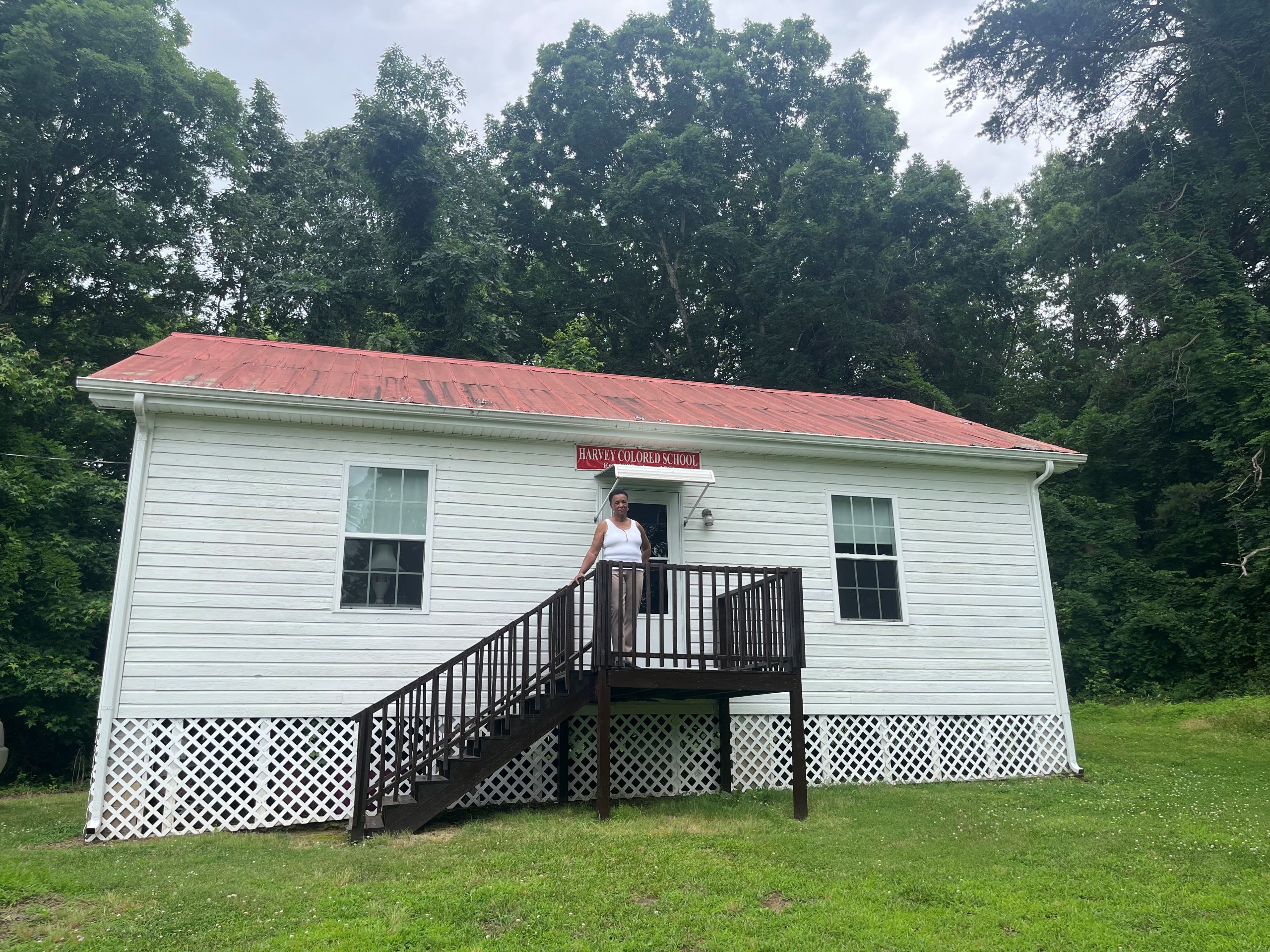 A woman, Annie Mosby, stands on the porch of a white one-room schoolhouse with a red tin roof. This is the Harvey School.
