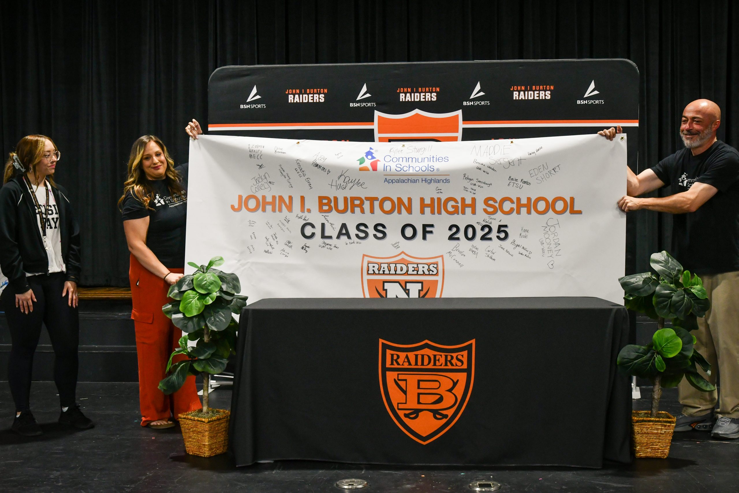 Three people in casual clothing stand on a stage with two holding a banner stating "John I. Burton High School Class of 2025." The banner is covered in signatures of students with notes about their plans for after high school.