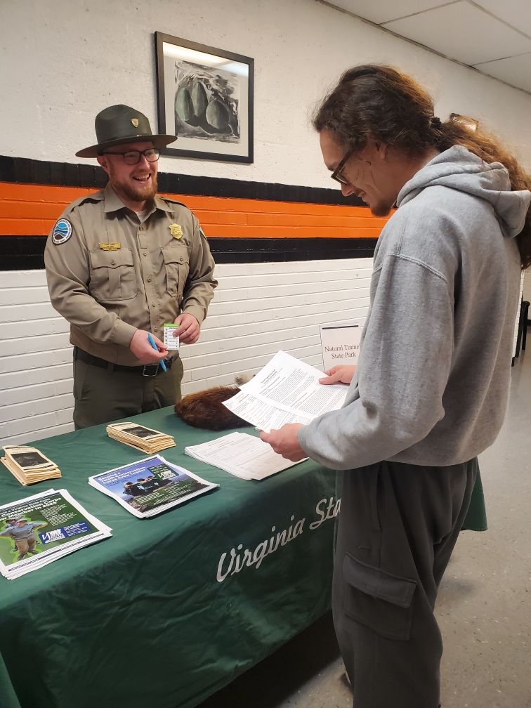 A smiling man in a Virginia State Parks uniform stands behind a table containing flyers and brochures about career opportunities with the park system. A student stands in front of the table, reviewing a sheaf of papers.