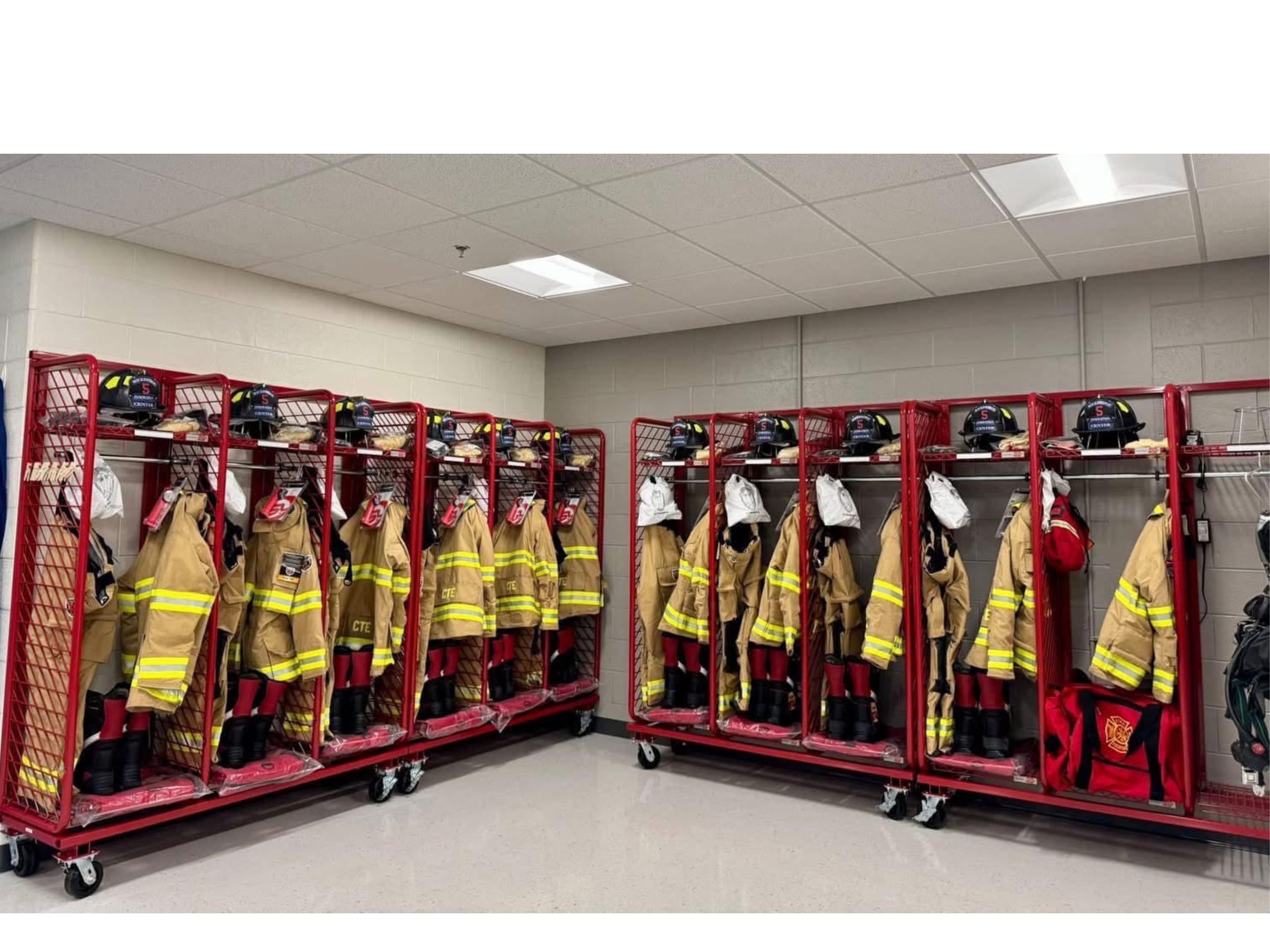 Two sets of lockers are shown in the corner of a room. Each red metal locker contains a reflective jacket, a black helmet, boots, pants, and a gear bag.