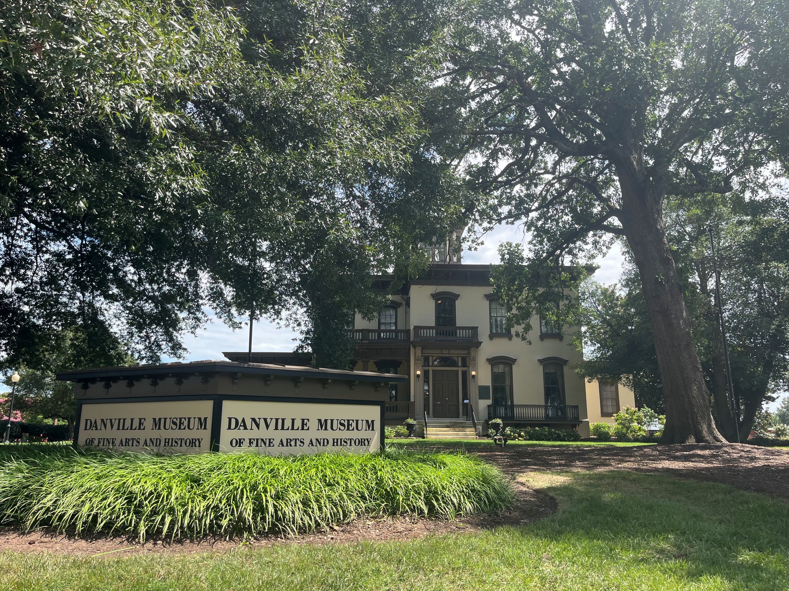 an exterior shot of the Danville Museum of Fine Arts and History, a tan and brown mansion, and a sign out front, surrounded by trees and landscaping