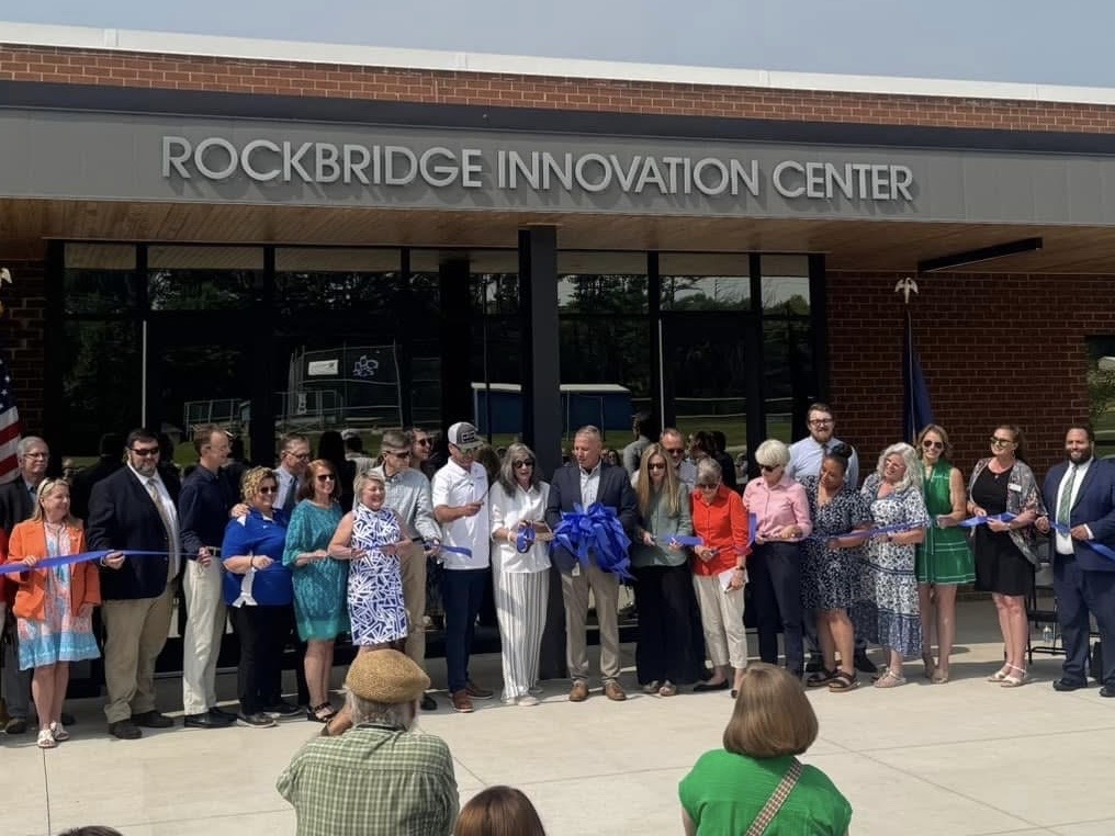 A line of people holds a large blue ribbon with a bow in the center on a sunny day. The building behind them has "Rockbridge Innovation Center" lettered on it.