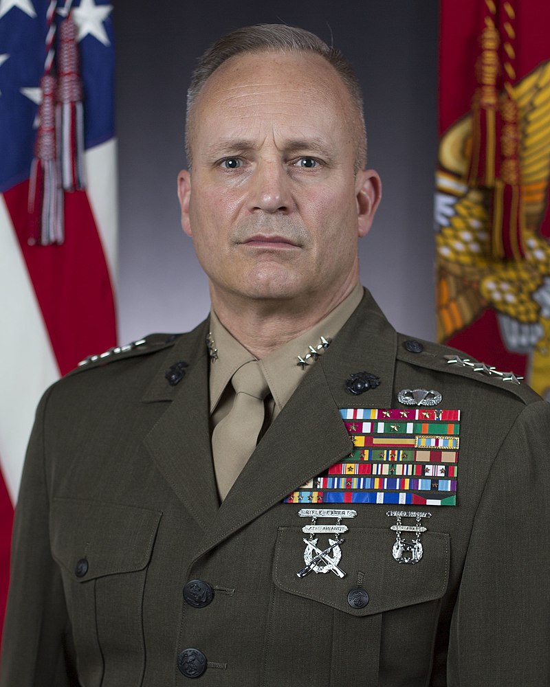 A man with close cropped hair wears a decorated military uniform in front of U.S. and Marine Corps flags.