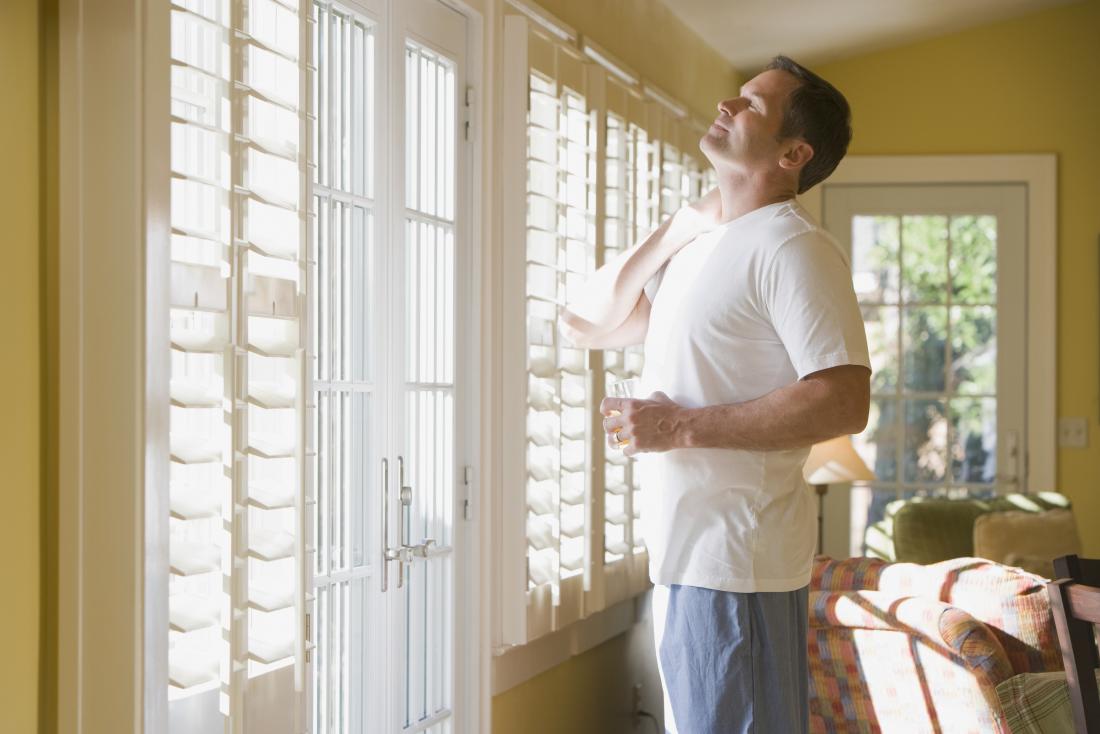 Man standing in house looking out of window having just woken up, holding stiff neck and glass of water.