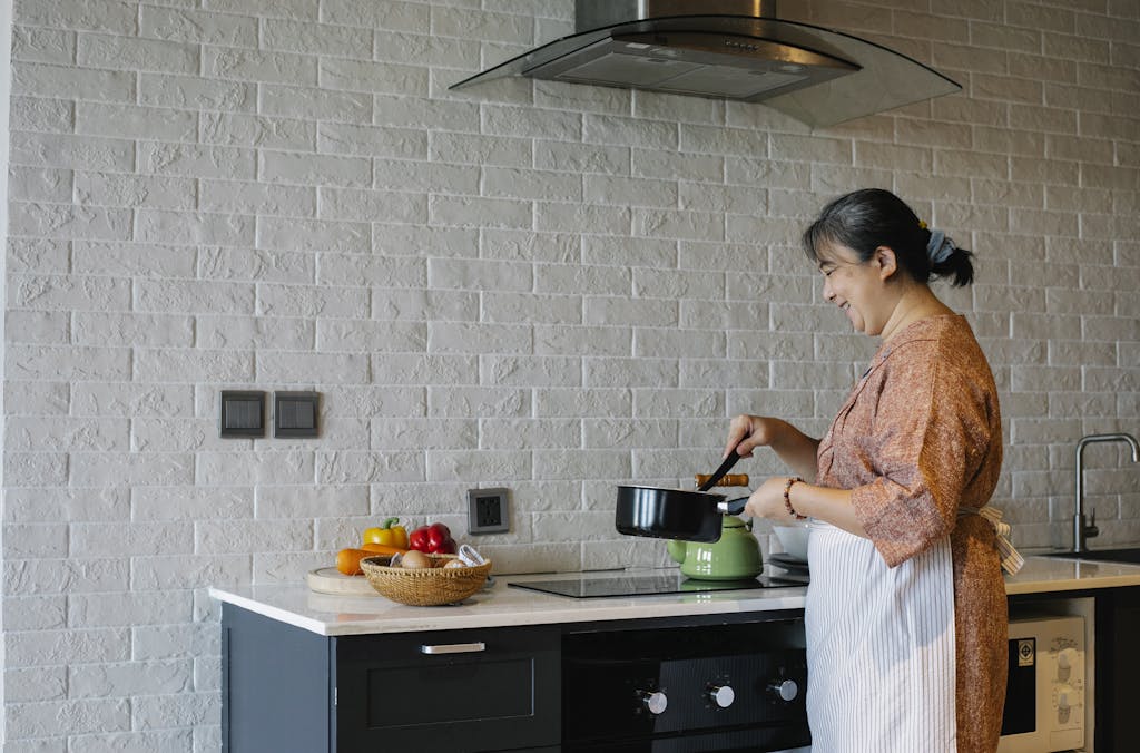 Side view of elderly female in apron standing near stove and holding pan while preparing lunch at home
