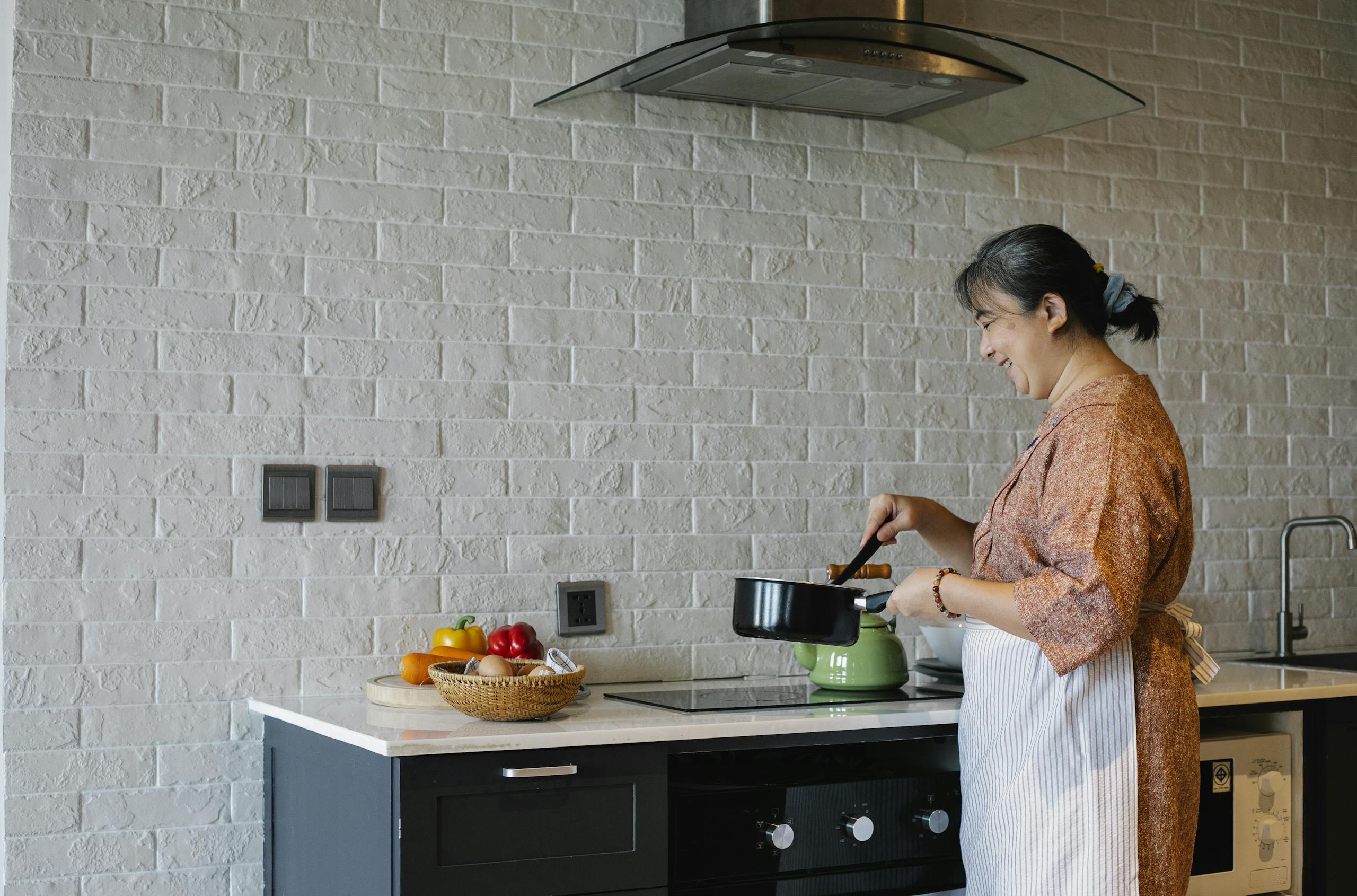 Side view of elderly female in apron standing near stove and holding pan while preparing lunch at home