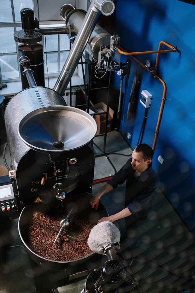 A man operates a coffee roasting machine in an industrial setting, surrounded by pipes and equipment.