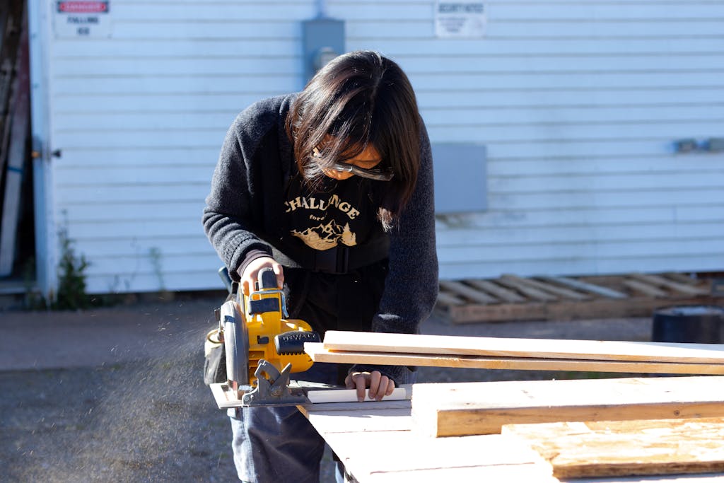 A woman using a cordless circular saw to cut wooden boards outdoors, emphasizing safety and precision.