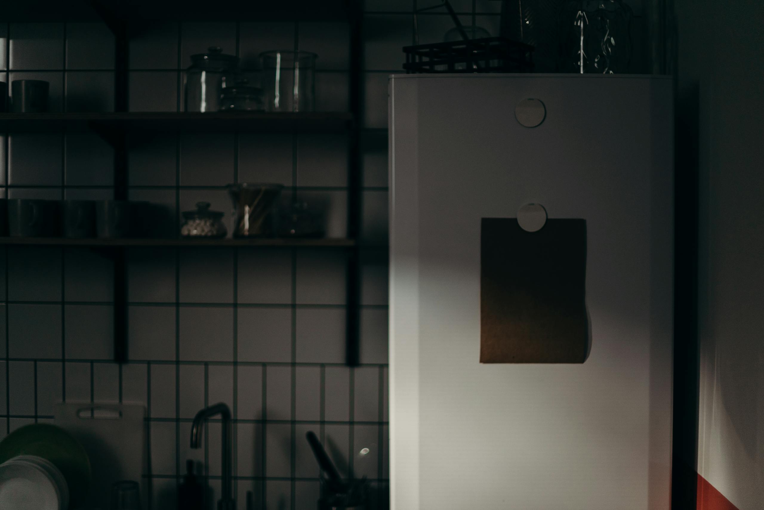 Dark and moody kitchen interior with fridge and shelves casting shadows, creating a minimalist ambiance.