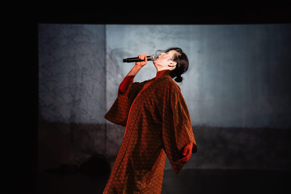 A woman stands center in profile, facing to the left of the photo. She wears a long rust-colored brocade jacket. Her head is thrown back and she holds a microphone to her mouth.