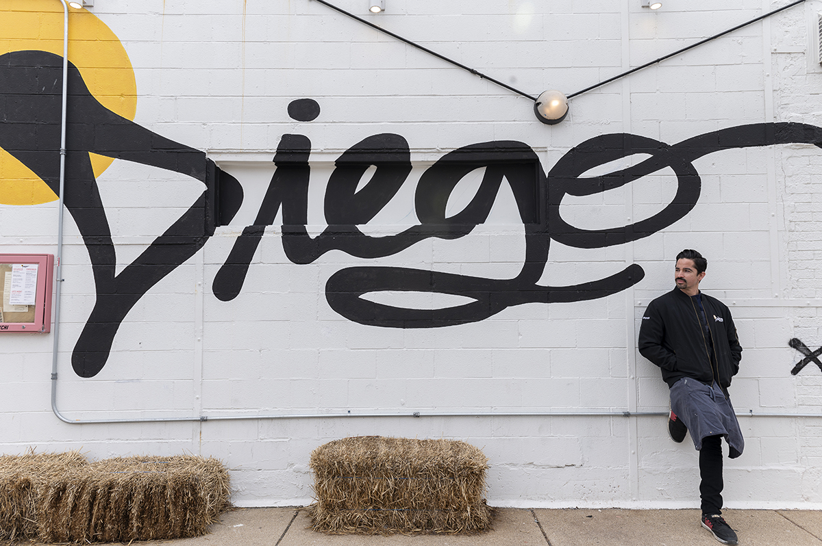 a chef leans against a white exterior wall with Diego painted in graffiti script