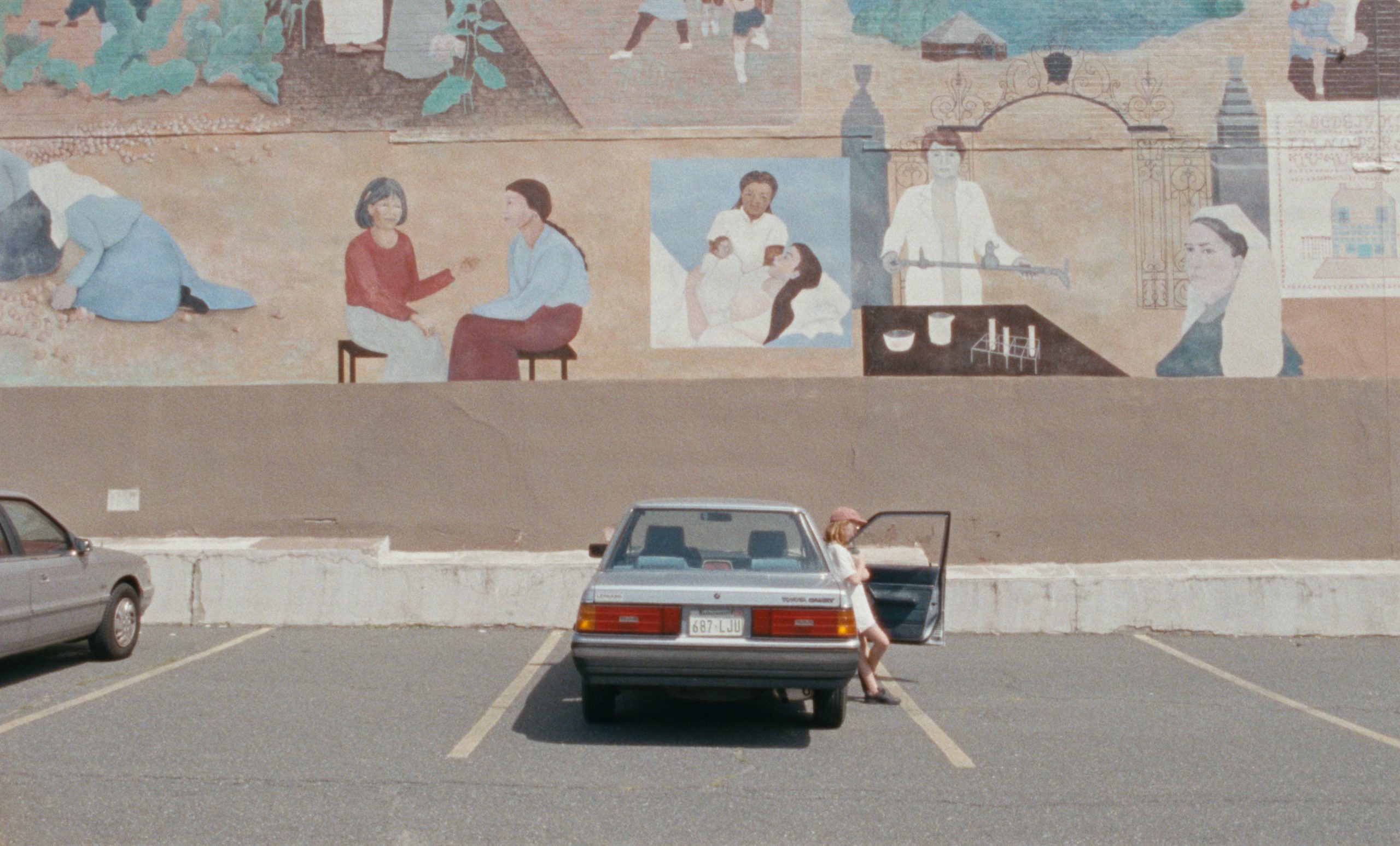 a young girl leans against a parked car in a mostly empty lot, with a mural in the background