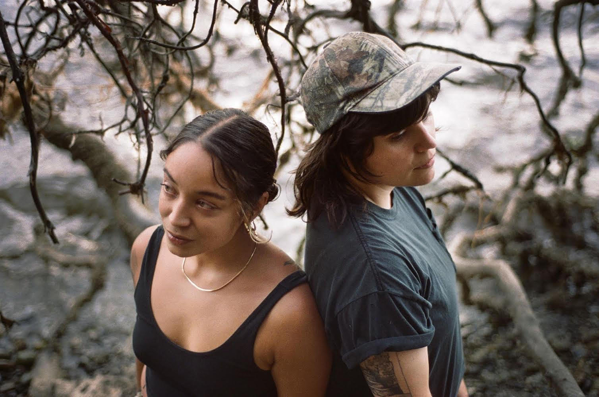 Two people stand back to back in front of branches and water