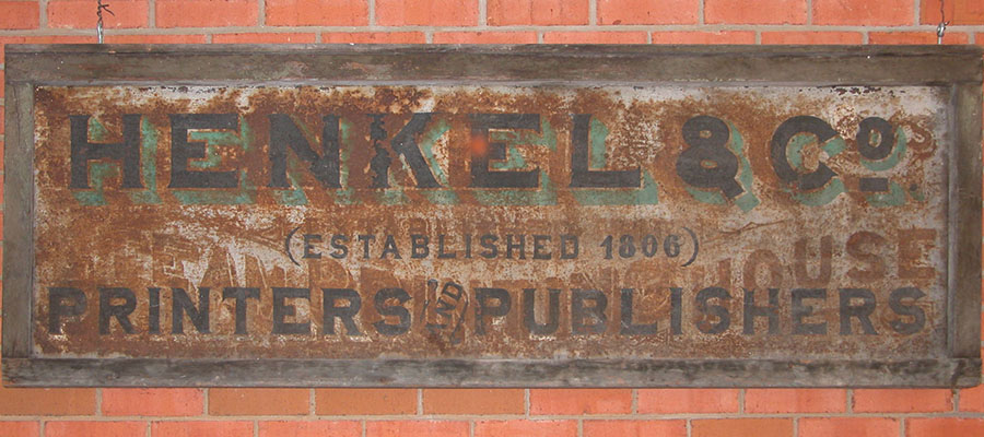 Modern color photograph of a Metal sign in a wood frame hanging in front of a brick wall reading: Henkel & Co. Printers and Publishers (established 1806).