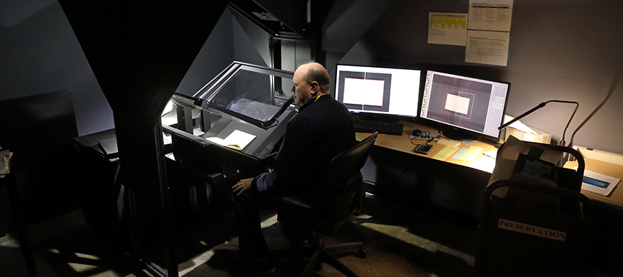 A white man sits in a darkly-lit room next to monitors showing the book he is scanning on a large format scanner