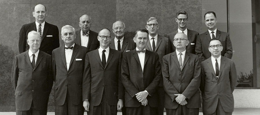 A black and white photo of 12 White men in suits standing in two rows outside the National Library of Medicine