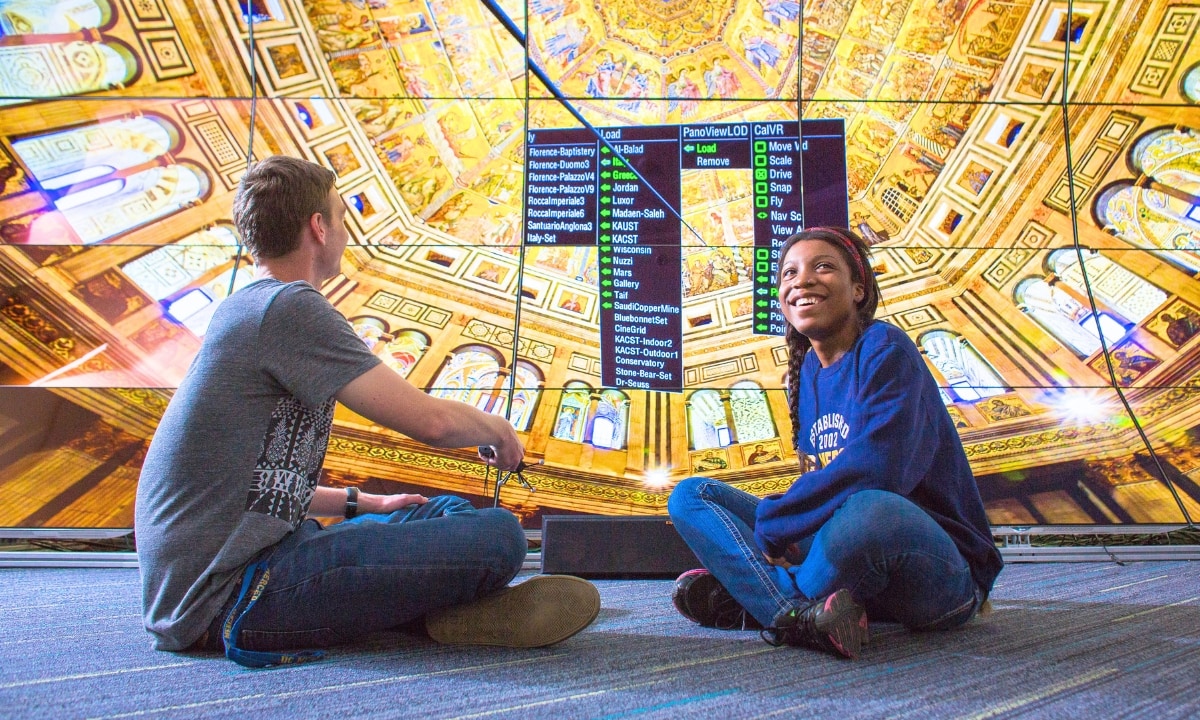 Two students in casual clothes seated with legs crossed on carpet in front of a large bank of lighted screens.