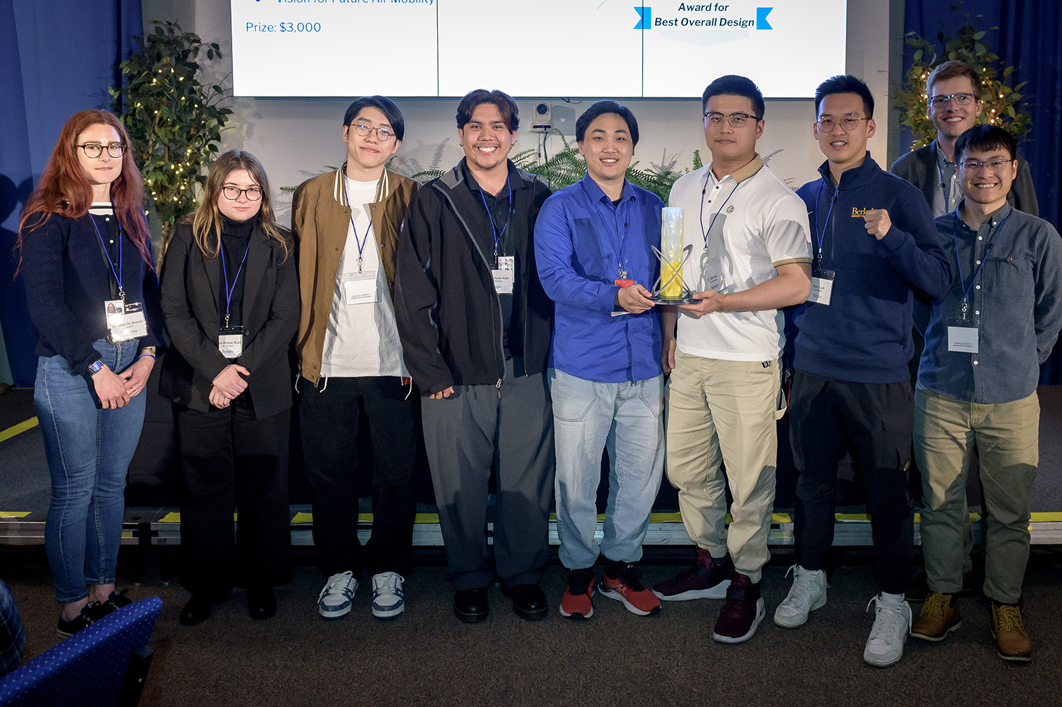 Nine people stand in front of a small stage. Two people at the center are holding a small trophy.