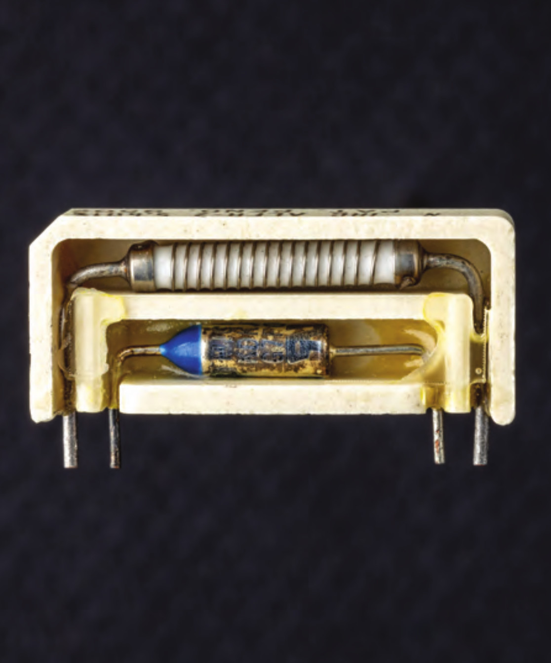 A photo from 'Open Circuits,' depicting a thermal fuse in cross-section. It is an oblong horizontal rectangle of off-white resin containing two compartments. The larger outer compartment contains a spring-wound white cylinder. The inner compartment is smaller and contains a glass resistor packed with small wires. Four metal leads depend from the casing.