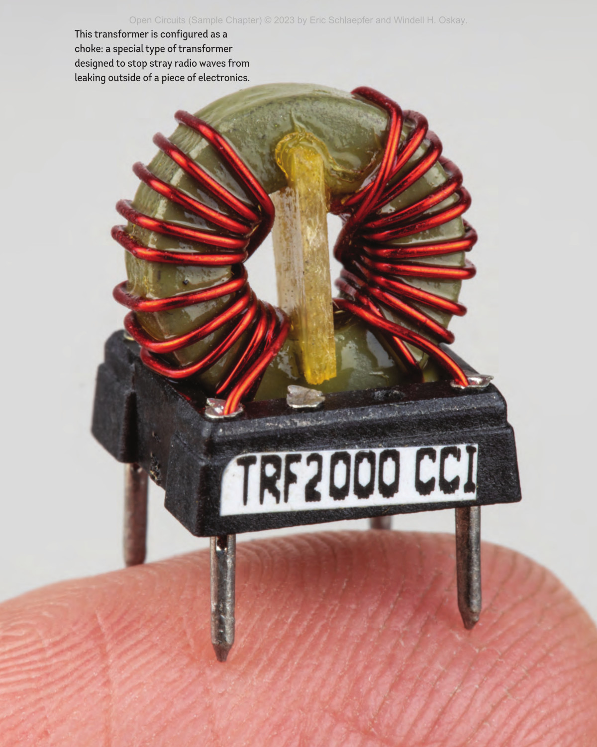 A photo from 'Open Circuits,' depicting a tiny toroidal transformer, wound with copper and mounted on a package for attachment to a circuit board, balancing on a man's fingertip.