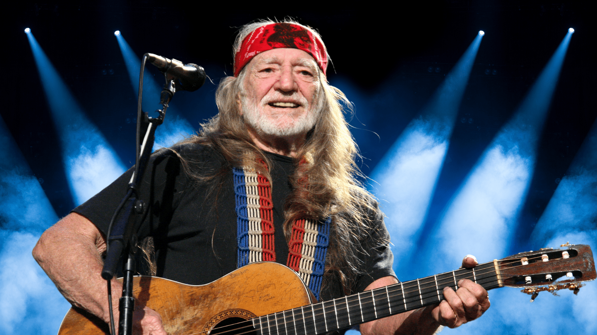 Willie Nelson holding a guitar and smiling on stage