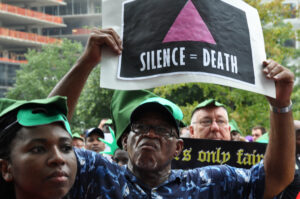Black man at march holding a sign. The sign has a pink triangle with the words "Silence = Death" in white underneath.