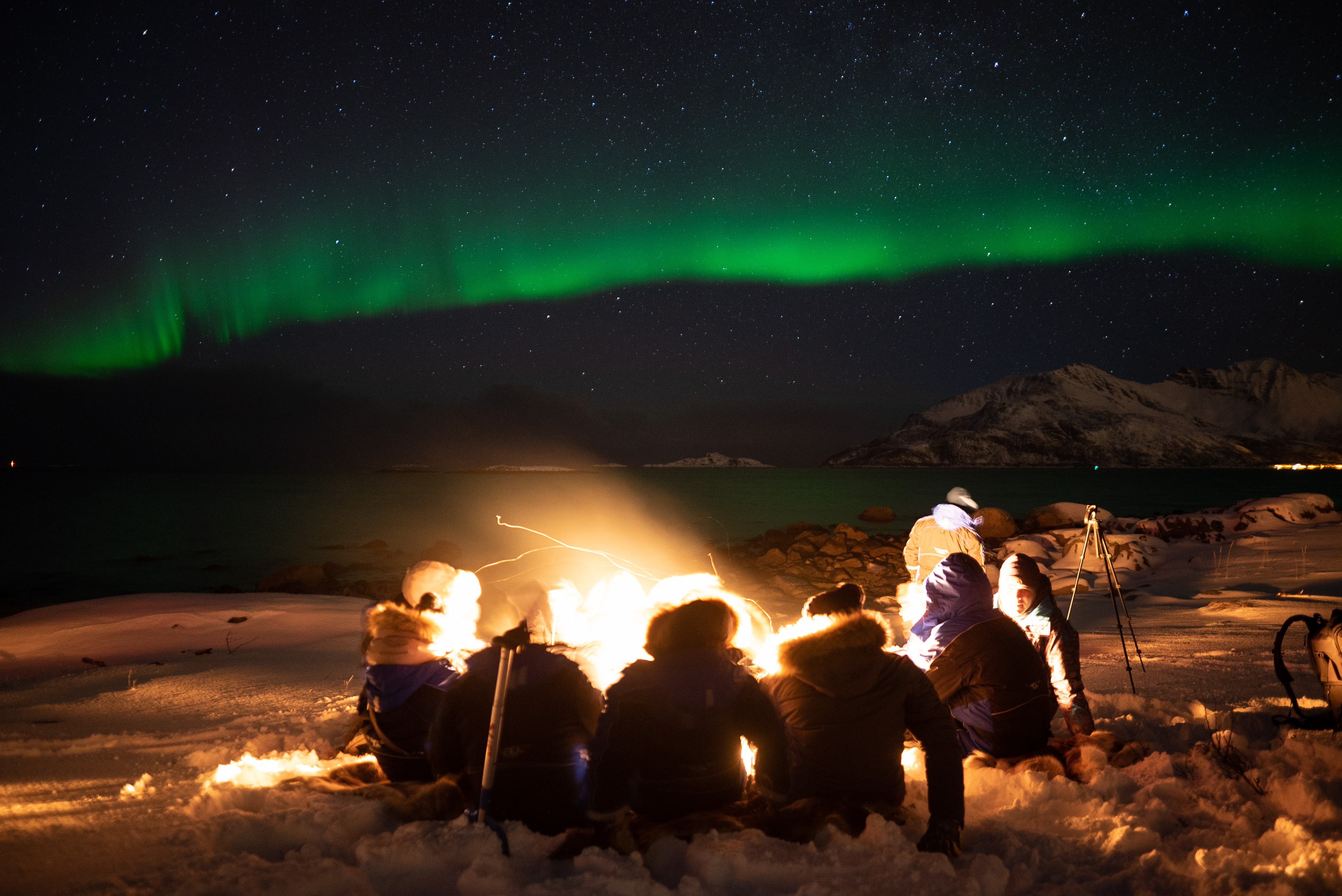 campfire in Norway surrounded with people wearing heavy coats and northern lights in the night sky