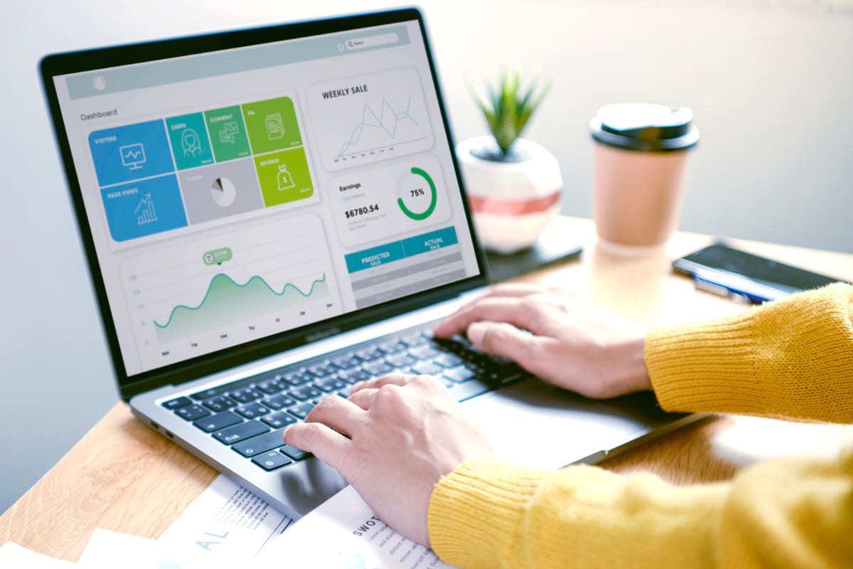 Google Tag Manager: como ele funciona e seu papel na estratégia de marketing? pessoa com casaco amarelo sentada em frente a uma mesa usando o notebook. Nesse último, estão sendo mostrados diversos gráficos, representando uma análise estratégica