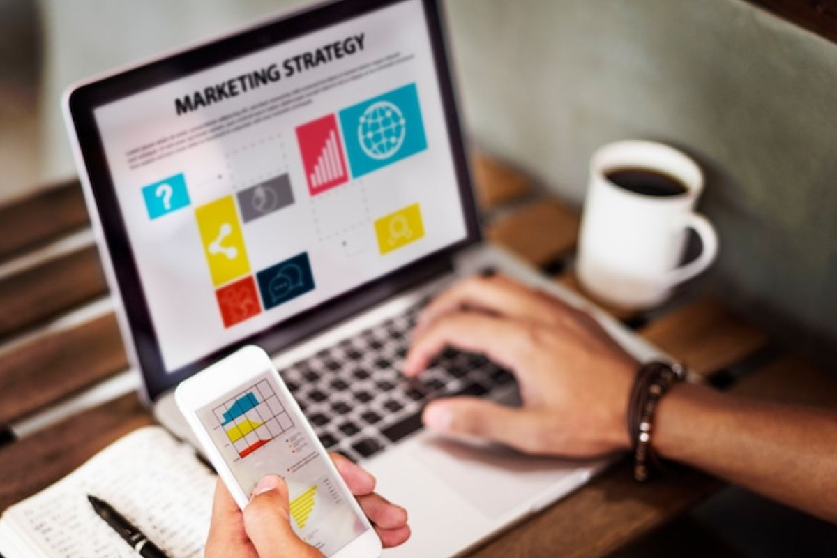 homem usando o notebook e o celular: marketing promocional homem sentado usando o notebook e o celular ao mesmo tempo. Ao seu lado tem uma xícara de café e um bloco de anotações