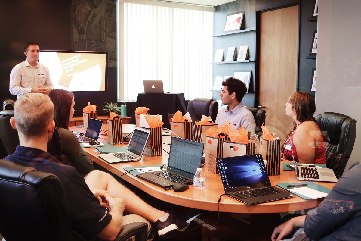 seis pessoas em uma sala de reunião, com uma mesa recheada de sacolas e laptops. A frente está um homem fazendo uma apresentação para os demais
