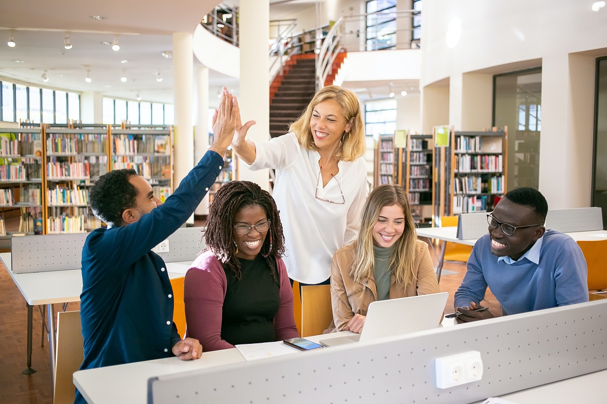 pessoas em reunião: automação de marketing pessoas diferentes sentadas em uma mesa de biblioteca, em reunião e sorrindo. Duas delas estão fazendo o sinal de high five