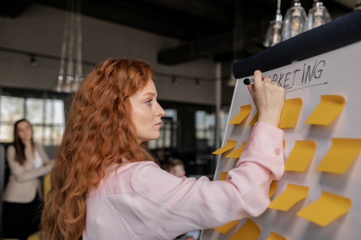 mulher escrevendo em quadro: plano de marketing mulher ruiva em pé, escrevendo em um quadro cheio de post-its