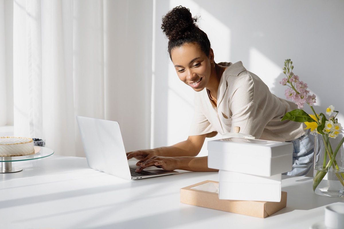 mulher usando o laptop: branding mulher negra de pé usando seu laptop em cima da mesa. Nessa última há flores e diversas caixas pequenas