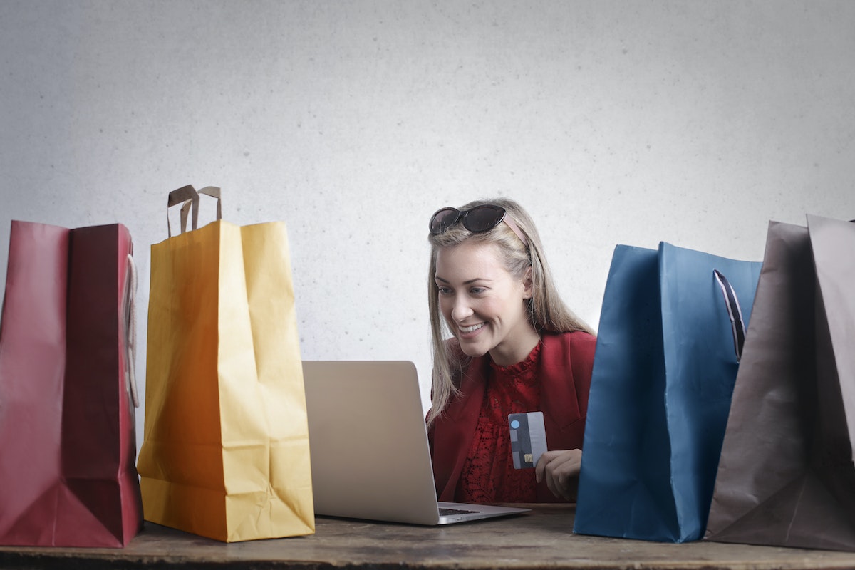 mulher loira, sentada em uma mesa cheia de sacolas. Ela está sorrindo para o laptop enquanto segura um cartão de crédito nas mãos, aparentando estar fazendo compras