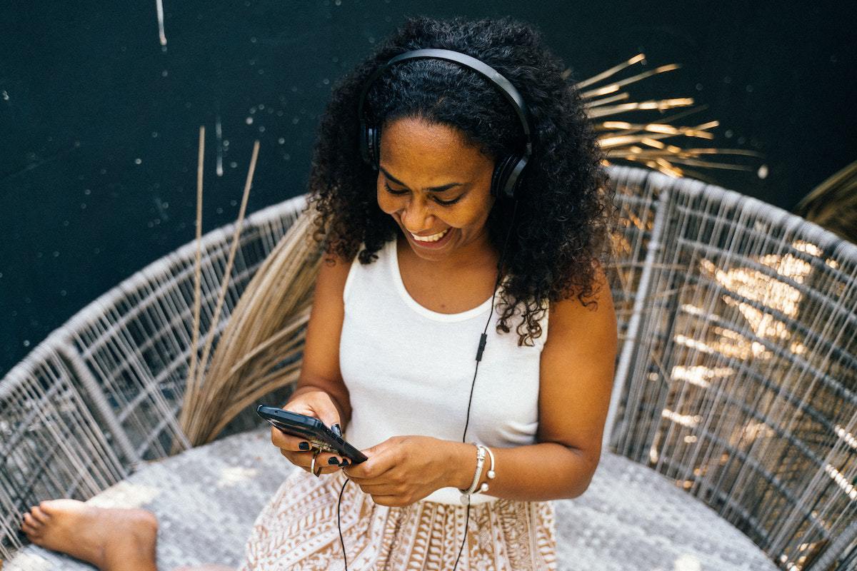 mulher ouvindo áudio no celular: podcasts mulher negrada sentada, usando fones de ouvido e segurando o celular com as mãos