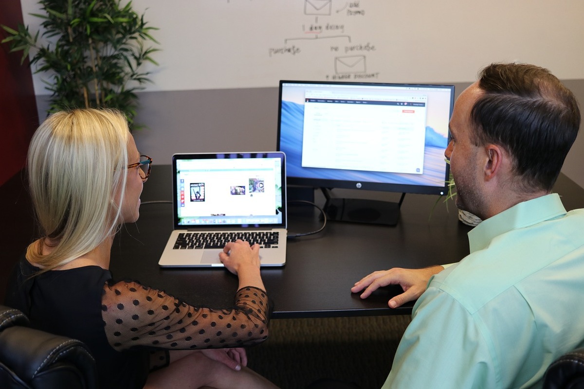 homem e mulher sentados com um laptop e um desktop à sua frente, analisando alguns dados