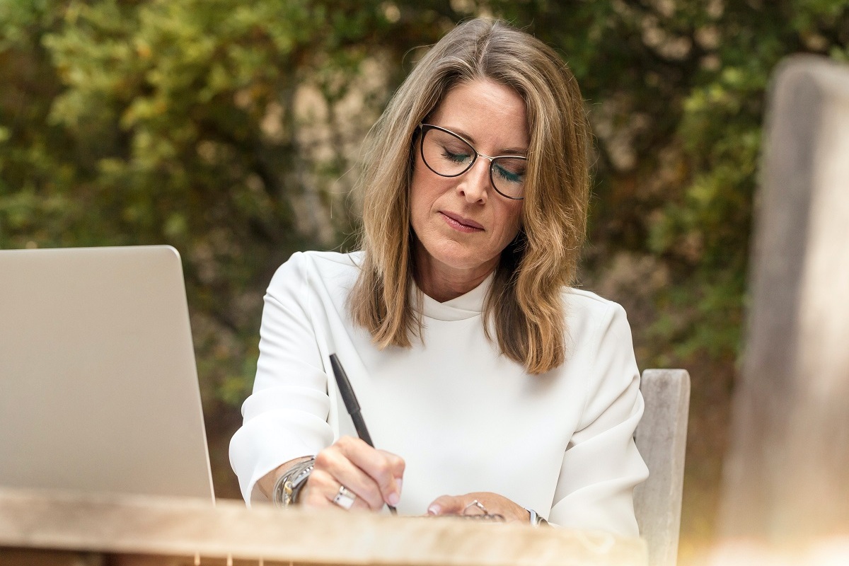 mulher loira escrevendo: guest post mulher loira sentada, com um laptop a sua frente e escrevendo algo em seu caderno