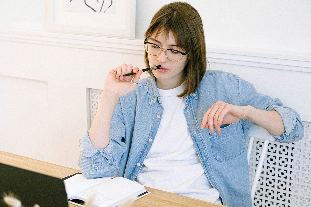 mulher sentada em frente a um laptop: guest post mulher asiática sentada com uma caneta na boa, olhando para sua mesa com um bloco de notas e um laptop