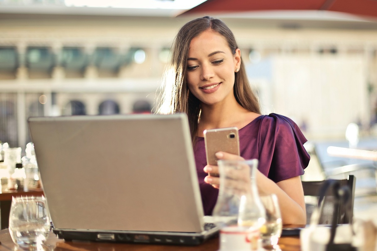mulher usando laptop e celular: twitter ads mulher sentada, sorrindo, usando o celular e o laptop ao mesmo tempo
